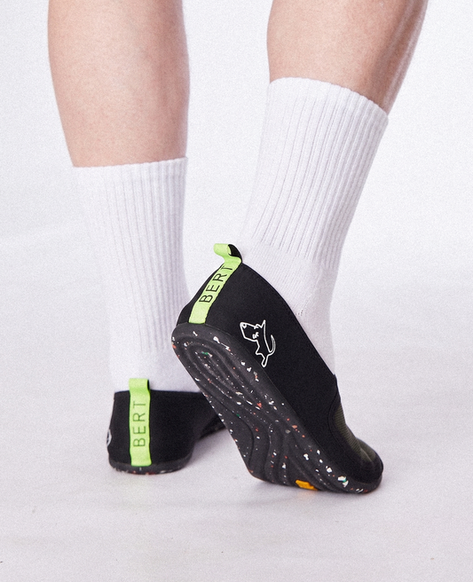 Black slip-on shoes with green accents, worn with white socks, featuring zero-drop soles and a dog logo.