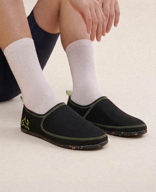 Black slip-on lightweight shoes with neon accents and white socks, worn by a seated person.