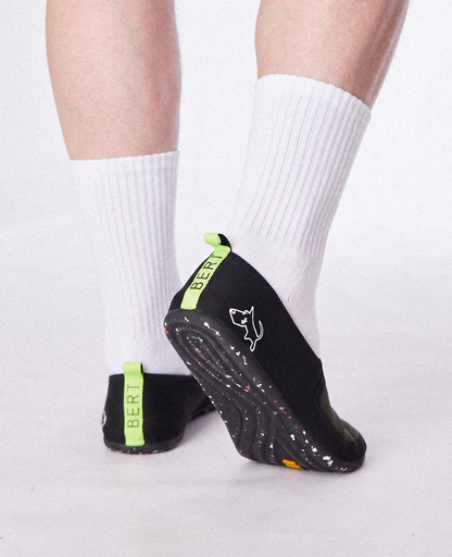 Black slip-on shoes with green accents, worn with white socks, featuring zero-drop soles and a dog logo.