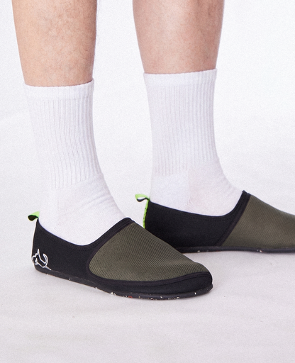 Person wearing olive green slip-on shoes with black trim and white socks on a white background.