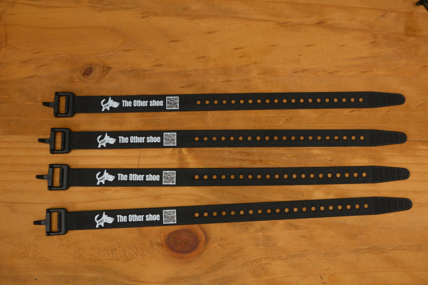 Four black adjustable shoe straps labeled 'The Other shoe' with QR codes on a wooden surface.