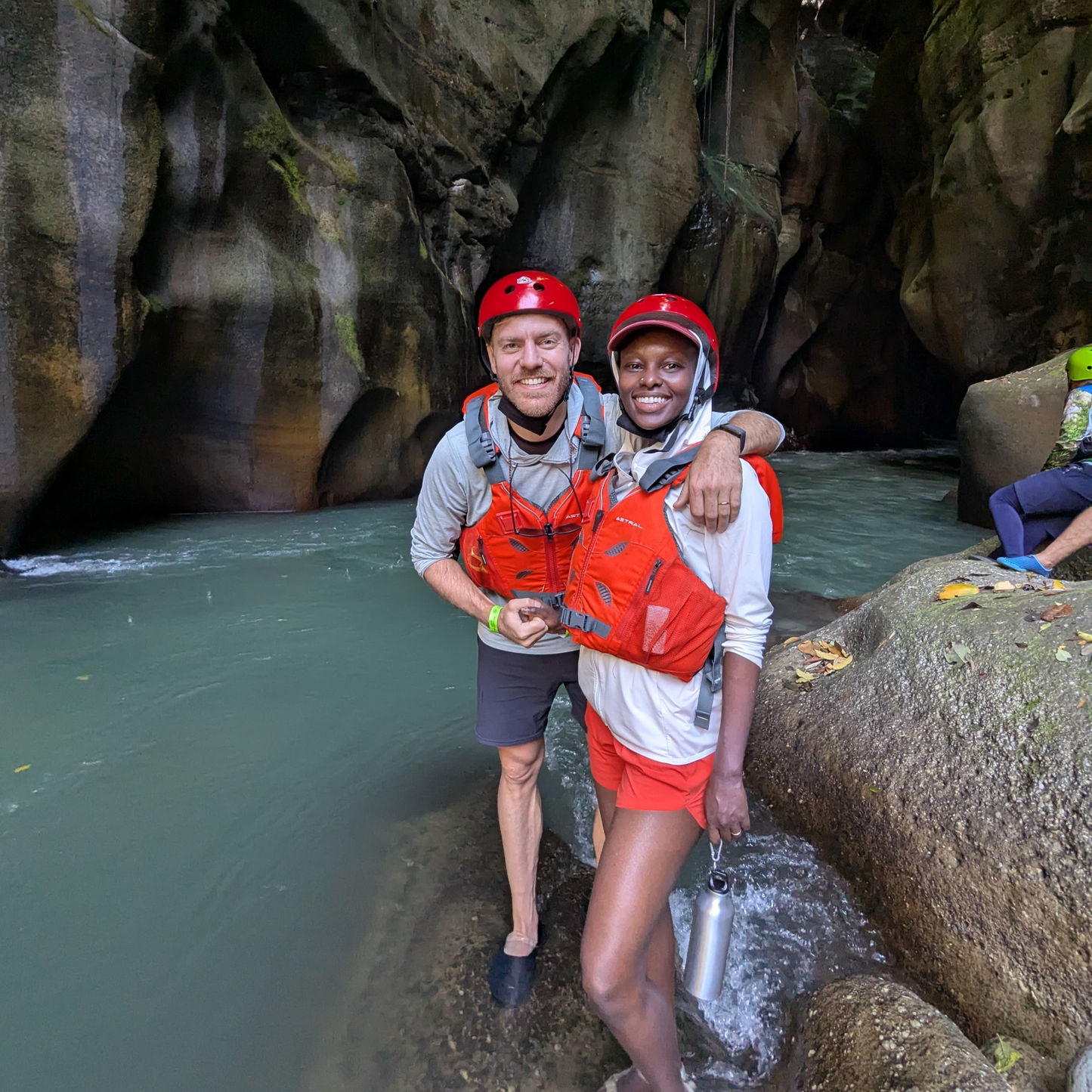 Two people wearing helmets and red life vests standing by river in rocky canyon, outdoor adventure