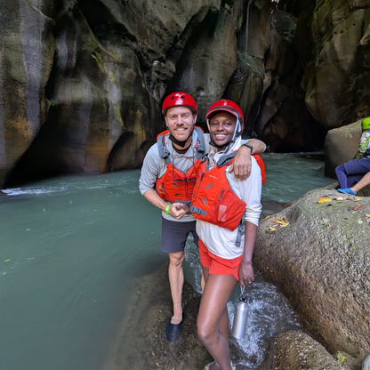 Two people wearing helmets and red life vests standing by river in rocky canyon, outdoor adventure