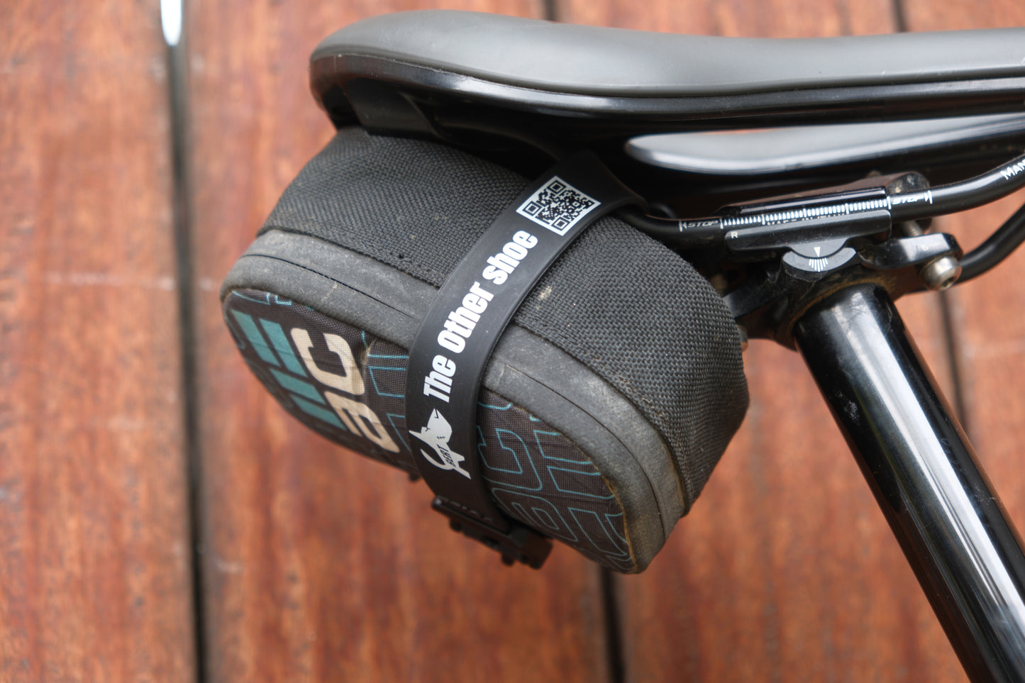 Black bicycle saddle with a compact black storage bag secured beneath it by a strap labeled 'The Other Shoe'.