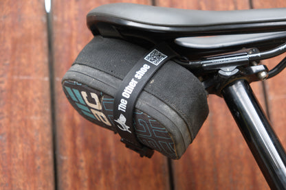 Black bicycle saddle with a compact black storage bag secured beneath it by a strap labeled 'The Other Shoe'.